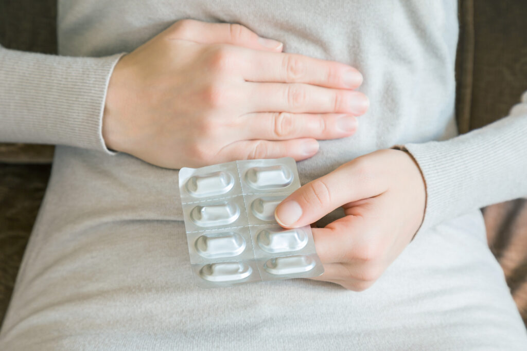 座っている女性が腸の部分に片手を当て、もう片方の手で薬を持っている様子