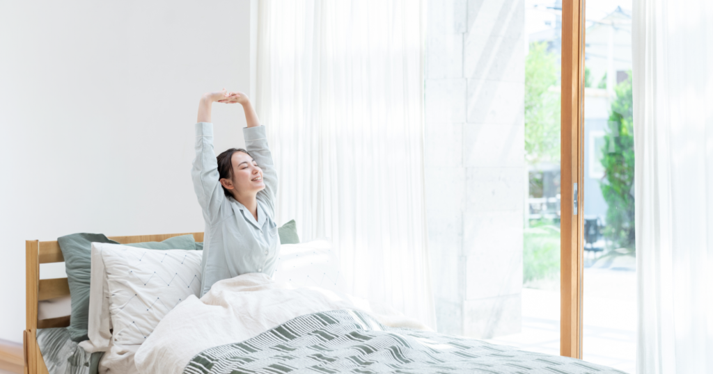 ベットに座った起きたばかりの女性が朝日を浴びて背伸びしている様子